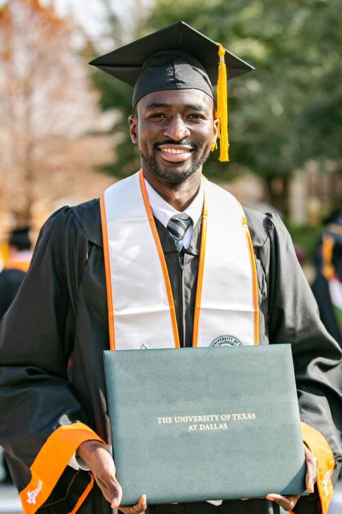 UTD graduate poses at commencement ceremony holding diploma cover. 